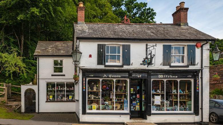 A white double fronted detached building with an old-fashioned shop front with large windows either side of the front door stands in front of tall green trees. The name A Coven of Witches is written in white on black woodwork above the windows.
