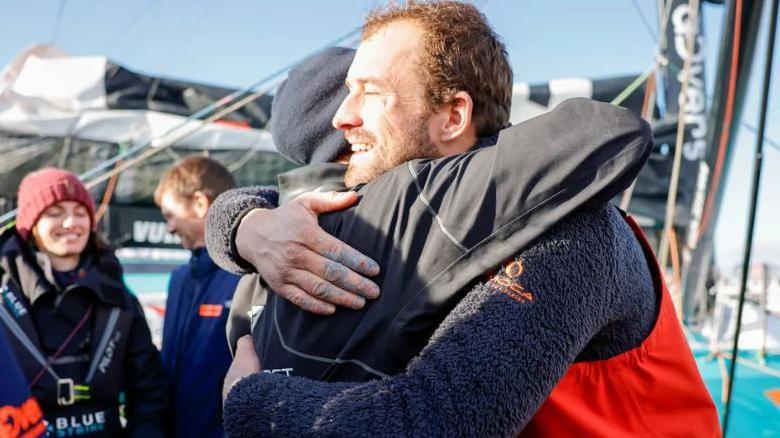 The back of a woman can be sent as she hugs a man in a sailing suit. Behind them two other people can be seen, including a woman with a woollen hat who is smiling. 