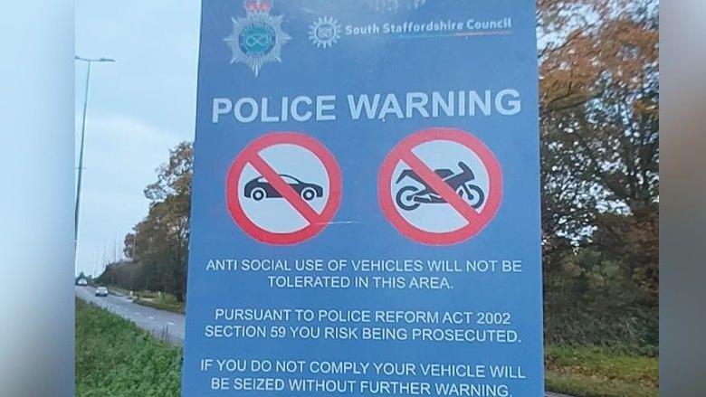 A blue rectangular road sign carrying a police warning stating anti social use of vehicles will not be tolerated. There are two circles with red borders, black symbols of a car and motorbike in each with a black diagonal line passing through them.