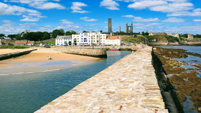 Harbour wall, with water either side, heading into the town of St Andrews, with historic buildings in the distance.