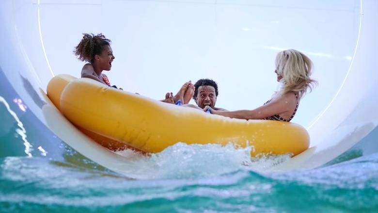 Three people in a giant floatable device come down a water flume and are about to splash into a swimming pool