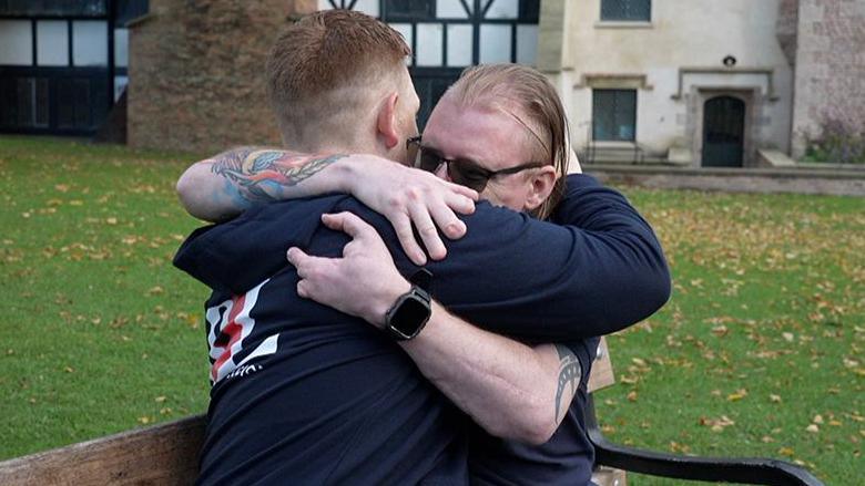 David Jack Plant and Antony “Coops” Cooper embrace on a bench outside a Tudor looking building