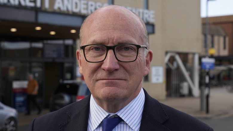 Nick Hulme smiles at the camera outside Colchester Hospital. He wears a black suit with a white shirt underneath that has blue stripes on it. He also wears a navy tie.