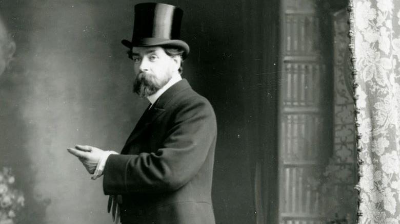 A black and white formal portrait photograph of Archibald Knox, who has a beard and is wearing a formal jacket and shirt with a stiff collar along with a top hat.