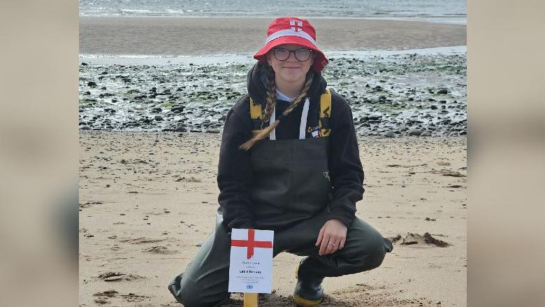 Lexie wearing red hat and water proof clothes while posing on the beach behind an England entry placard.