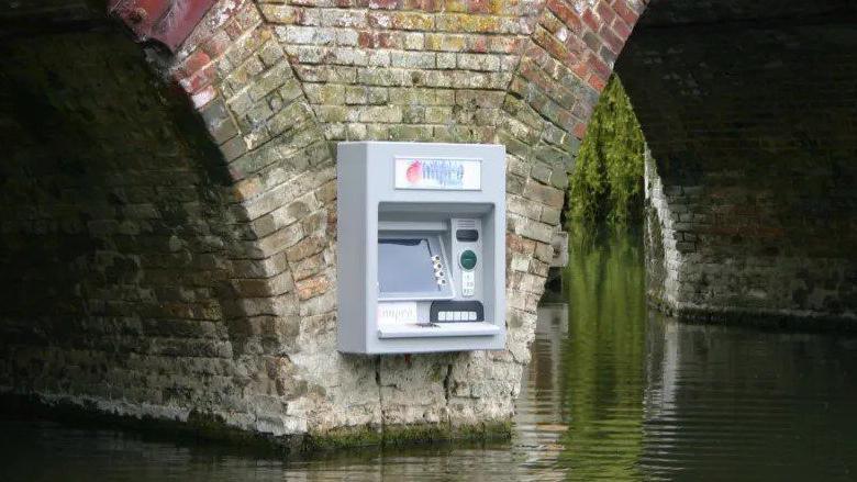 Facade of grey atm machine with screen, buttons on brick buttress with rippling water below 