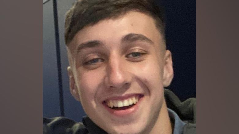 A young man smiles at the camera in a close-up shot. He has short dark hair.