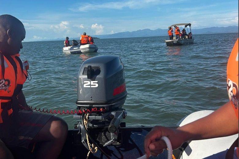 An image taken from near the back of a power boat showing the engine of the boat at the back with two people wearing orange life jackets operating the boat. The view looks out on to the blue sea with two other boats on the water with people wearing orange life jackets in them. There are mountains in the distance and white clouds in the blue sky