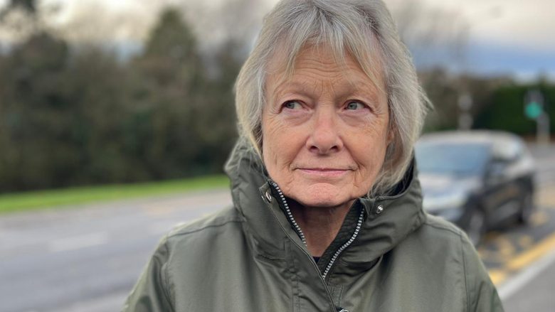 A woman on the side of the road, behind her is a black car driving, she is the focus of the image. She is looking at the camera, she has grey hair and is wearing a khaki cagoule. 