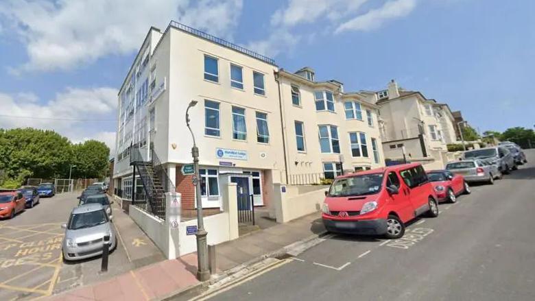 A Google Street image of the outside of a blocky, beige building with a number of cars parked outside on the street.