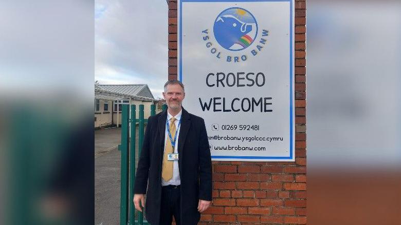 Peter Evans, with short hair and a grey short beard, standing in front of a sign welcoming visitors to Ysgol Bro Banw school, wearing a long black coat, yellow tie and lanyard