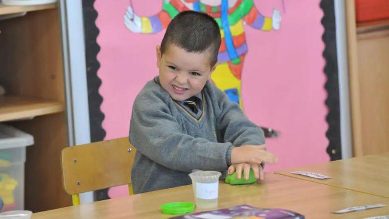 A young child in a school uniform stands next to a table. He is playing with a green item that looks like clay.