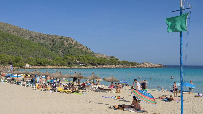 A sandy beach with sunbathers, swimmers, a green flag on a pole and straw beach umbrellas. A rocky spur is in the background at the far end of a bay.