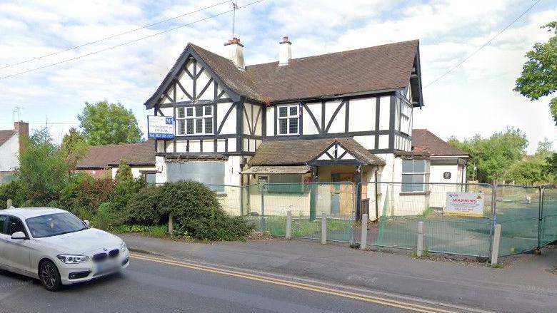 The shell of the fire-damaged pub, a mock-tudor black and white building dating back to the 1920s. Metal security fencing surrounds the site with a safety warning notice attached.