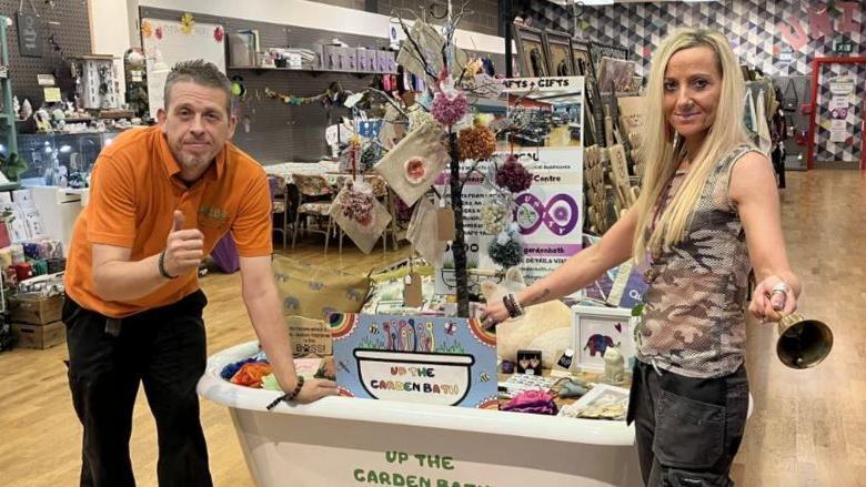 Dave - a man with light brown hair, wearing an orange T-shirt and black trouser - standing next to a white bathtub inside the Unity store with Kez - a woman wearing a printed sleeveless shirt and cargo trousers, standing on the other side of the bathtub, holding a bell.