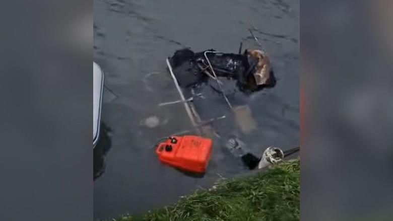 A pile of burnt debris, including an orange jerry can and what is left of a gutted canal boat, on a canal, near the grass bank.