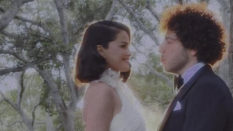 Selena Gomez, a young woman with dark hair, wears a sleeveless, high-necked white wedding gown and is embracing Benny Blanco, a young man wearing a dark tuxedo and white shirt. They are standing in front of trees.