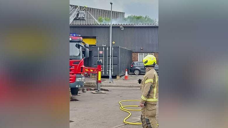 Smoke is seen rising from an industrial building as a firefighter in uniform watches on. There is a hose on the ground beside him.