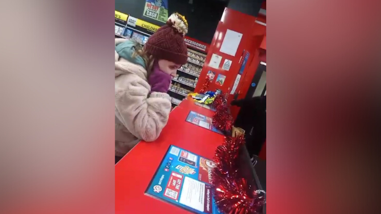 A teenage girl is covering her ears with her fingers. She wearing gloves, a hat and a cost. She's standing at the cash register in a shop. Tinsel is decorating the counter top. Shelves of DVDs are in the background.