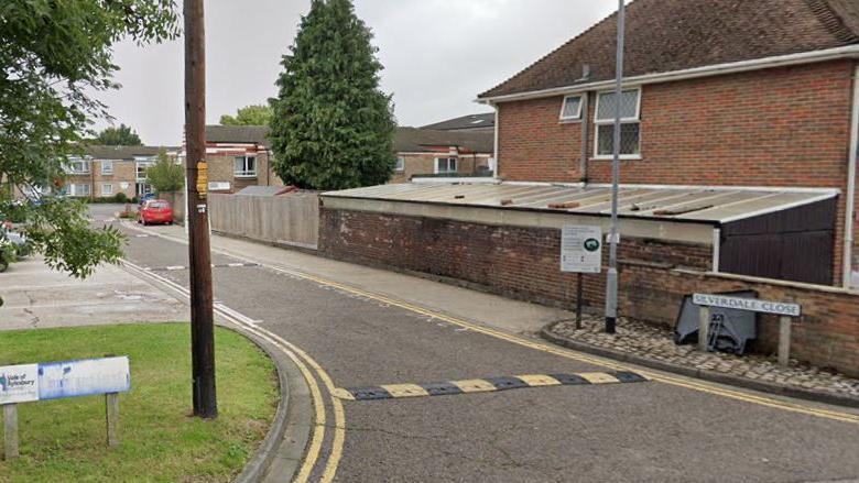 Silverdale Close, which has yellow and black speed humps and small blocks of flats. There is a tall conifer tree and a small red car in the distance. 