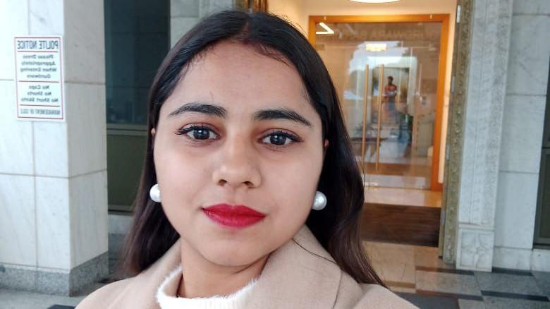 Harshita Brella looks into the camera for a selfie while standing outside a building. Her hair is open, and she is wearing bright red lipstick and pearl drop earrings.