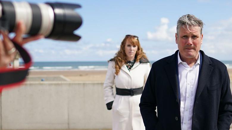 Labour Party leader Sir Keir Starmer and Angela Rayner, deputy leader and chairwoman of the Labour Party visit Seaton Carew seafront on 1 May 2021 in Seaton Carew. Sir Keir is in the foreground with his hands in his pockets, with Rayner behind him. A camera shoots a photo, with the beach behind them.