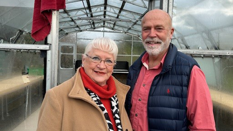 A woman with white hair and glasses in a brown coat and red scarf beside a man with a white beard, red shirt and blue gilet stood in front of a greenhouse