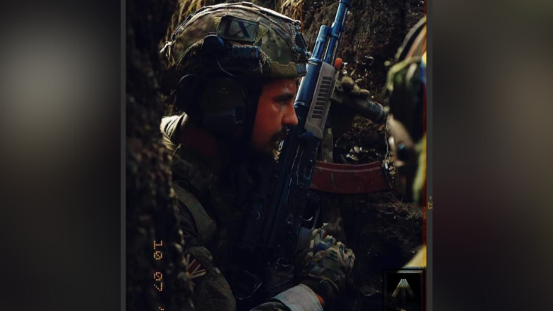 A soldier is wearing a helmet and gloves in a khaki pattern and is carrying a gun. He is standing in a dug out shelter with a wall of soil on both sides of him.