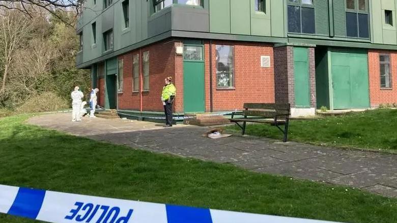 Two people in white forensic suits examine the ground while a police officer in uniform looks on.