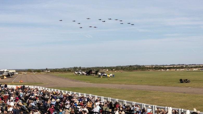 Crowds of people are on the ground behind a white fence, separating them from a runway and grassy area. High in the sky are 15 planes, so high they mostly look like black dots. The sky is a clear blue and they are getting into formation - flying closely together.