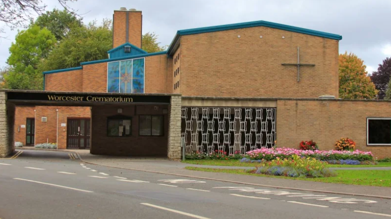 The exterior of Worcester crematorium, a brick building by the side of a road, next to a grassy patch with flowerbeds. The building has a chimney and a stained glass window is visible. A sign above the entrance reads "Worcester Crematorium".