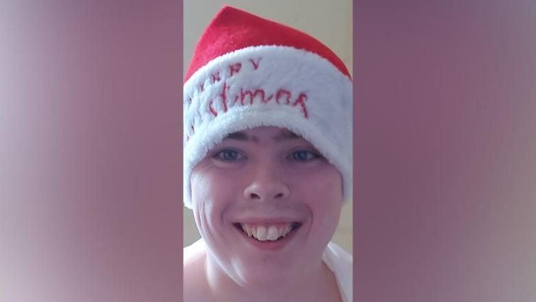 Liam Gunn looking at the camera
He is smiling and wearing a red hat with a white fur trim with Merry Christmas written on it