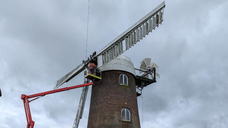 Under a dark grey sky, the top of the windmill, which is a round red brick building with a white dome on top, has a cherry picker lifting men to the white sails as they remove one