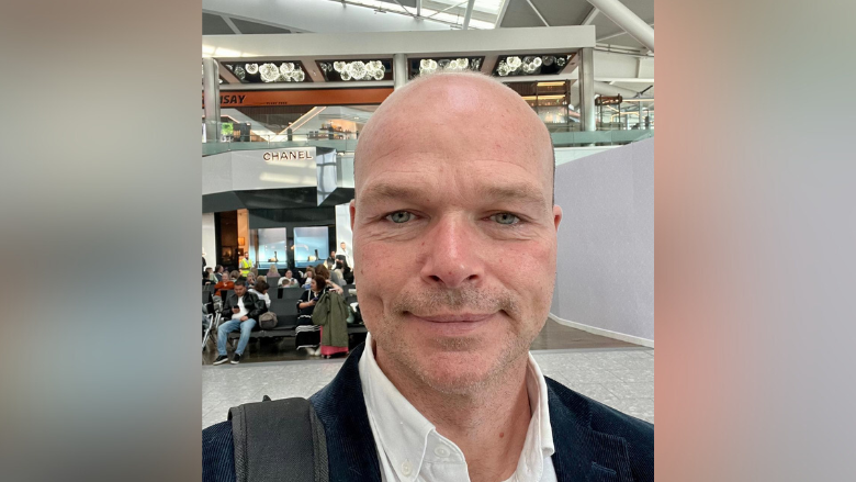Eamon Devlin smiles for a selfie. He has a shaved head and is wearing a smart white shirt with a blue blazer over top. A large black strap of a shoulder bag can be seen on his right shoulder. In the background people can be seen on seats, possibly at an airport.