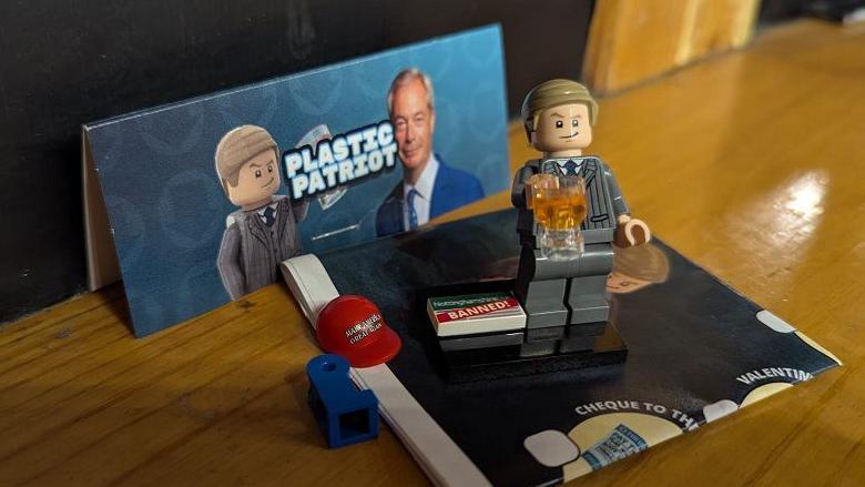 Nigel Farage Lego figure clutching a beer, against a backdrop of "plastic patriot" packaging