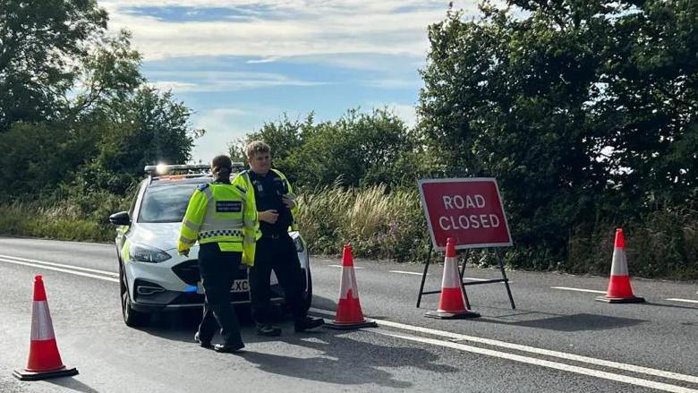 A police car and cones block the road, with a 'Road Closed' sign