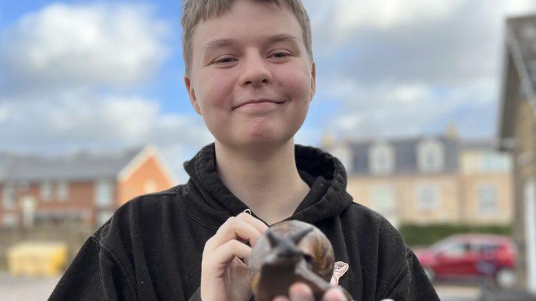 Connor Bouttell, wearing a black hoodie and holding a large snail in the palm of his hand. He has short brown hair and is smiling at the camera. The background, which is blurred, shows a residential scene with houses.