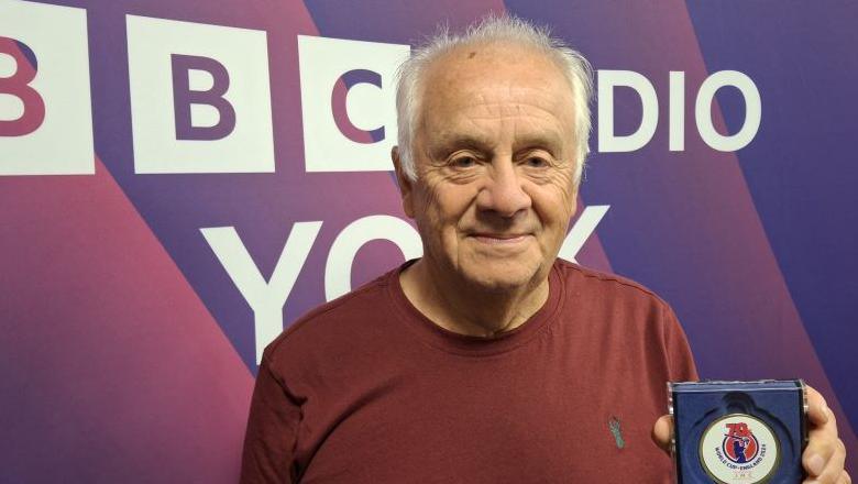 Mick Kenyon in the Radio York studio wearing a red t-shirt and holding up his trophy for winning the World Cup