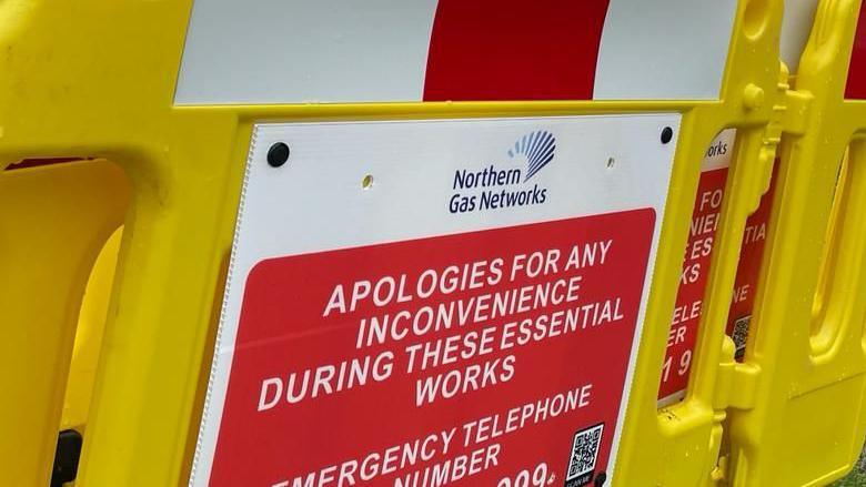 A red and white sign which reads "Apologies for any inconvenience during these essential works". The sign is stuck to a yellow road barrier and also features the logo for Northern Gas Networks.
