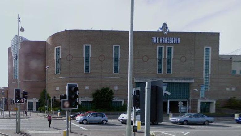 Two-storey brick building which curves round to the right in line with the road. It has tall narrow windows on the first floor and "THE HARLEQUIN" in 20s-style lettering towards the top above two of the windows. There are cars on the road in front of the building and a set of traffic light.