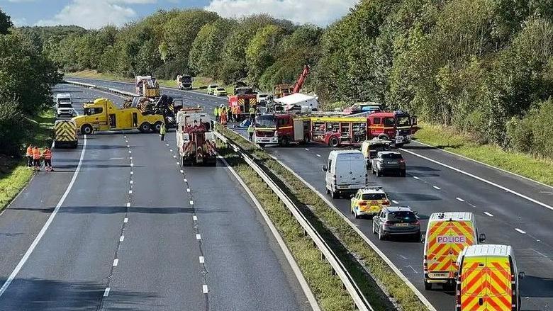 A view looking down the M4 from a bridge. At the front are vans with high visibility backs, there are various other cars and police cars. In the distance is a fire engine and a large yellow recovery vehicle.