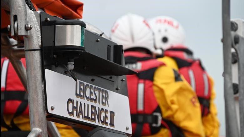 The rear of a lifeboat, the view consisting of a sign on a metal pole reading: Leicester Challenge III. To the right, and slightly out of focus there are two people in yellow lifeguard outfits with red safety vests and white helmets marked RNLI.