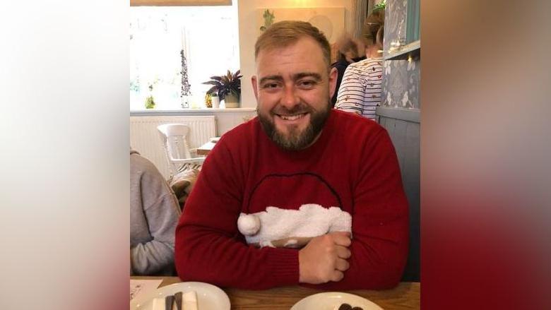 Andrew Darn pictured smiling and sitting at a table. He is wearing a red jumper with an image of Santa Claus.