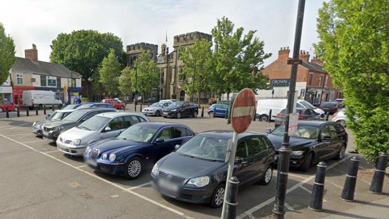 Google view of the Sheep Market car park in Spalding town centre. There are black bollards around the perimeter of the car park and a number of vehicles in bays. There are shops and other buildings in the background.