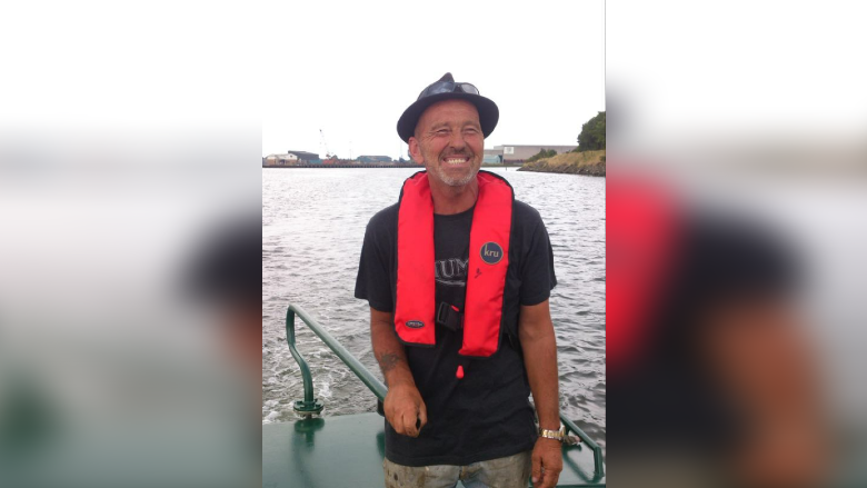 Graham Bennett smiles at the camera in a picture taken as he is sailing down a river. He is wearing a hat with sunglasses perched on top of it and has a life-preserving aid around his neck.