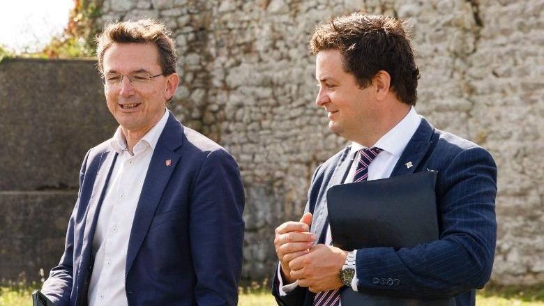 Two men in blue suits walk through a heritage site with old walls