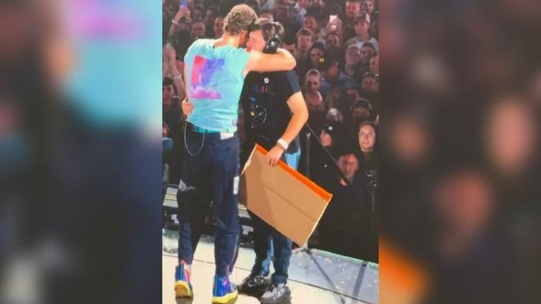 Chris Martin hugs AJ Murphy on stage, with the crowd behind them