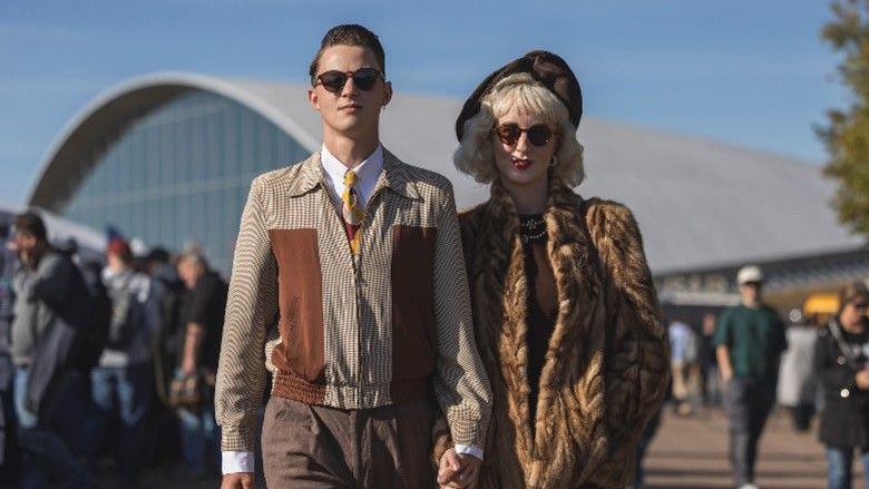 Visitors dressed in vintage attire at the 2024 Battle of Britain Air Show. The man and woman are holding hands and walking towards the camera with straight expressions. They are both wearing sunglasses and the woman has medium length curled hair and a fur coat. The man's hair has been slicked back and he is wearing a striped brown jacket over a white shirt and tie.