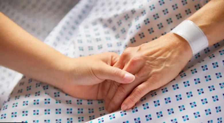 Two people hold hands on a medical bed. A younger hand clasps that of an older adult.