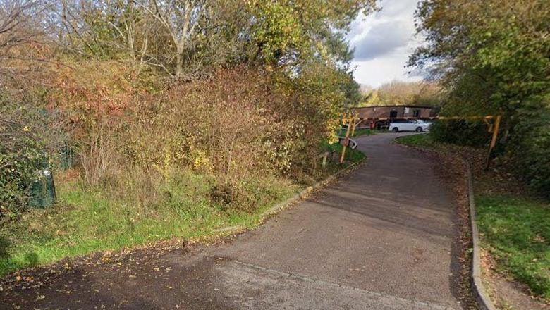 A narrow road with woodland on either side, which leads to a car park with a white car parked in a bay. 